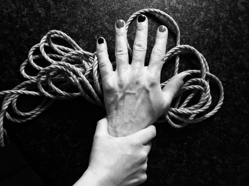 ein Bündel Seile, darauf liegend eine Hand mit lackierten Fingernägeln, die von einer anderen Hand gepackt wird